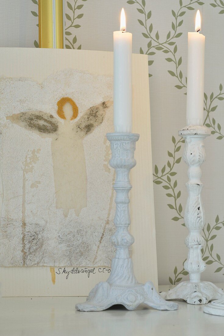 White candlesticks and picture of angel in front of wallpaper with pattern of delicate climbing plants