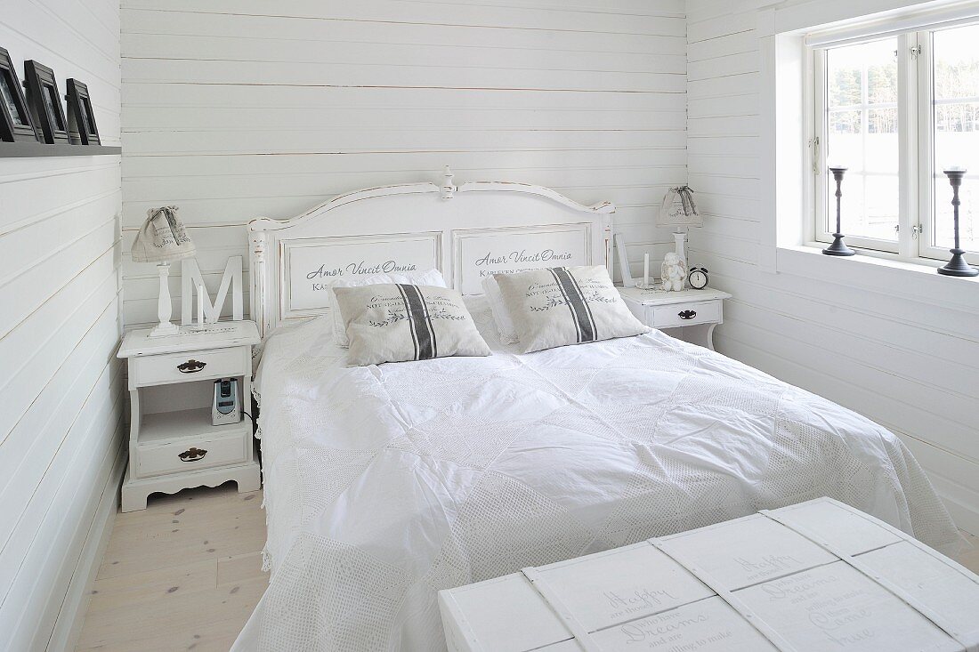 Romantisch dekoriertes Doppelbett und Nachtschränkchen im reinweissen Shabby Schlafraum eines skandinavischen Holzhauses