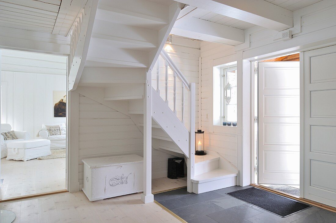 Foyer with winding wooden staircase and view into living room in wooden house painted entirely white and with white furnishings