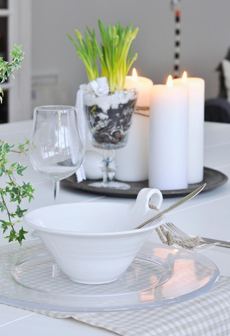 Henkelschale mit Schnurdeko, Glasteller und Weinglas als Gedeck auf Esstisch; Kerzen und Pflanzendeko auf Tablett im Hintergrund