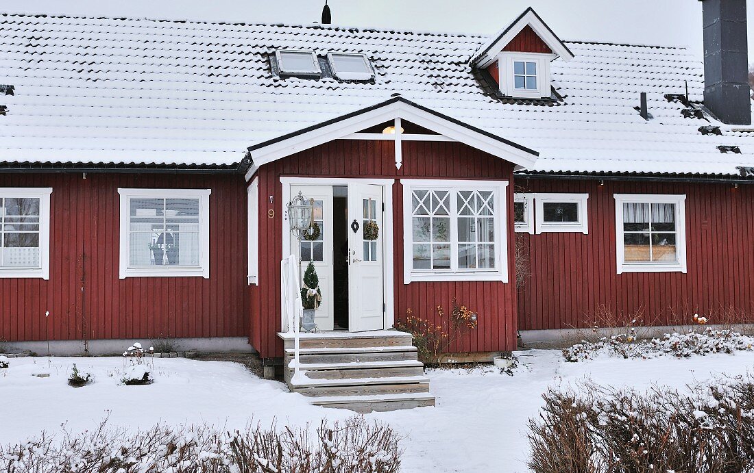 Verschneites, skandinavisches, Holzhaus mit roter Holzverschalung und geschmückter Tür am Eingangs-Vorbau
