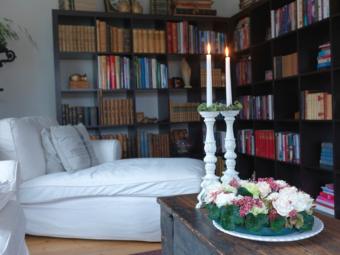 Wreath of flowers and lit candle in white candlesticks on trunk; white couch and L-shaped bookcase in background
