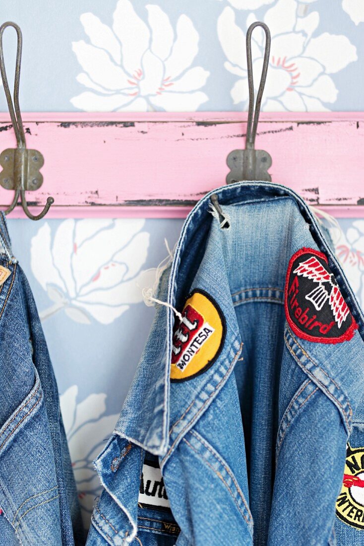 Mit Textilstickern dekorierte Jeansjacke an rosafarbener Vintage Hakenleiste an pastellblauer Blütentapete