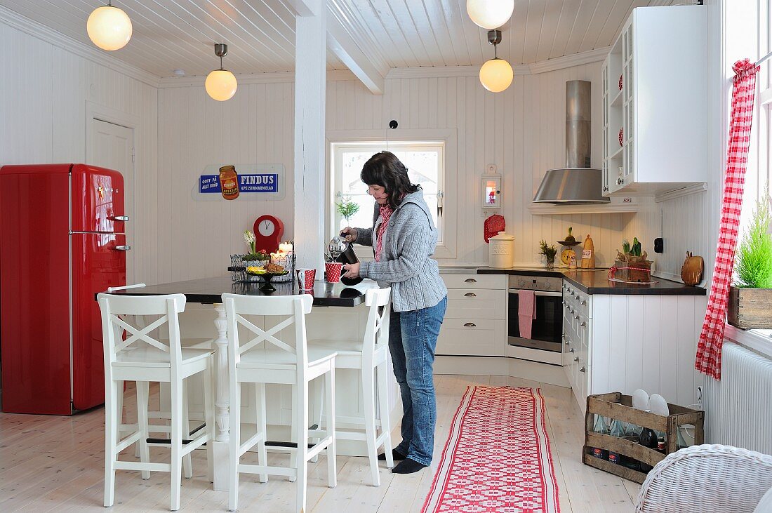White, Scandinavian, country-house kitchen with red, retro fridge and woman pouring drinks at breakfast bar