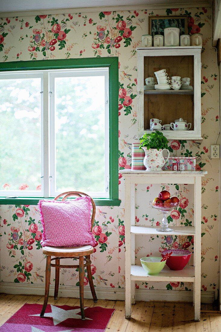 Thonetstuhl vor Fenster, daneben Beistelltisch und Hängeschränkchen an tapezierter Wand mit Blumenmuster