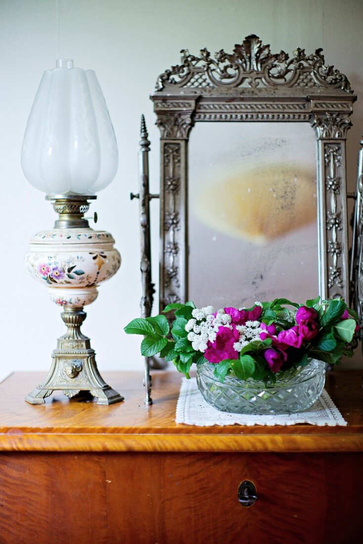 Paraffin lamp and bowl of flowers in front of vanity mirror with ornate silver frame on simple wooden chest of drawers