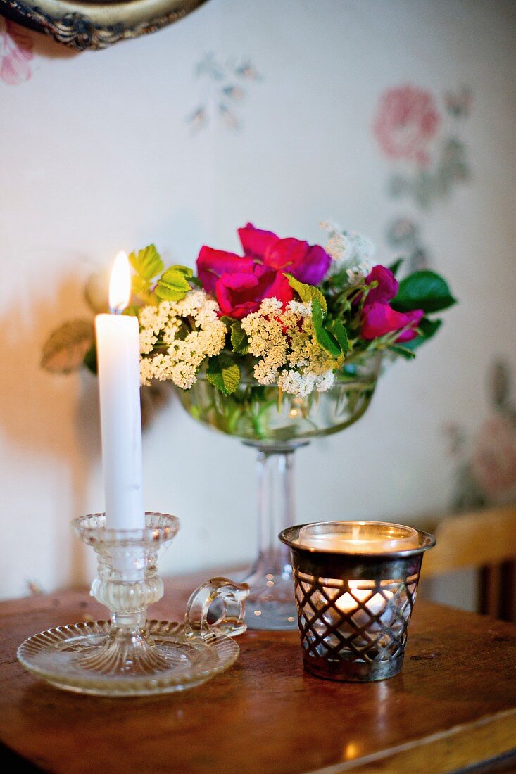 Vintage Glas Kerzenhalter mit brennender Kerze neben Windlicht, dahinter Glasschale mit Blumen