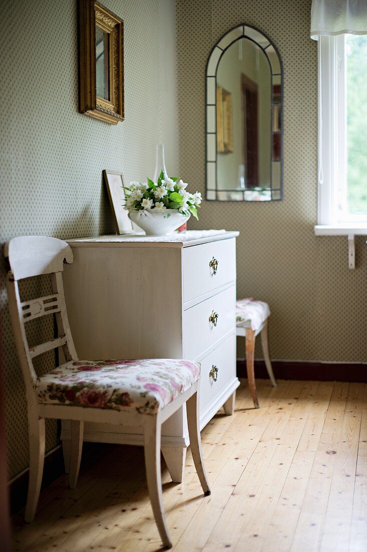 Upholstered, white-painted kitchen chairs flanking chest of drawers in rustic interior with well-tended wooden floor