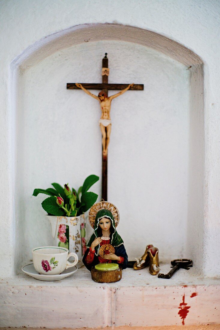 Devotional objects in small niche; statue of saint next to teacup and flowers in jug and crucifix on wall