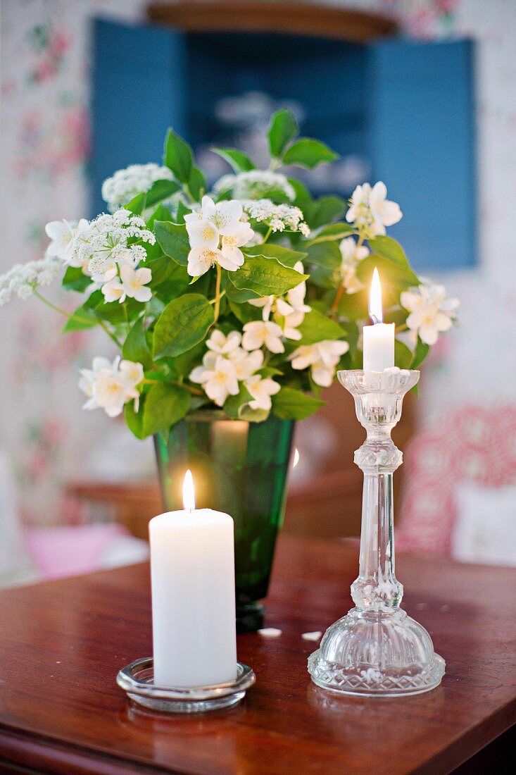 Stumpenkerze und Kerze in Glas Kerzenhalter vor Jasminblüten in grüner Vase auf Holztisch