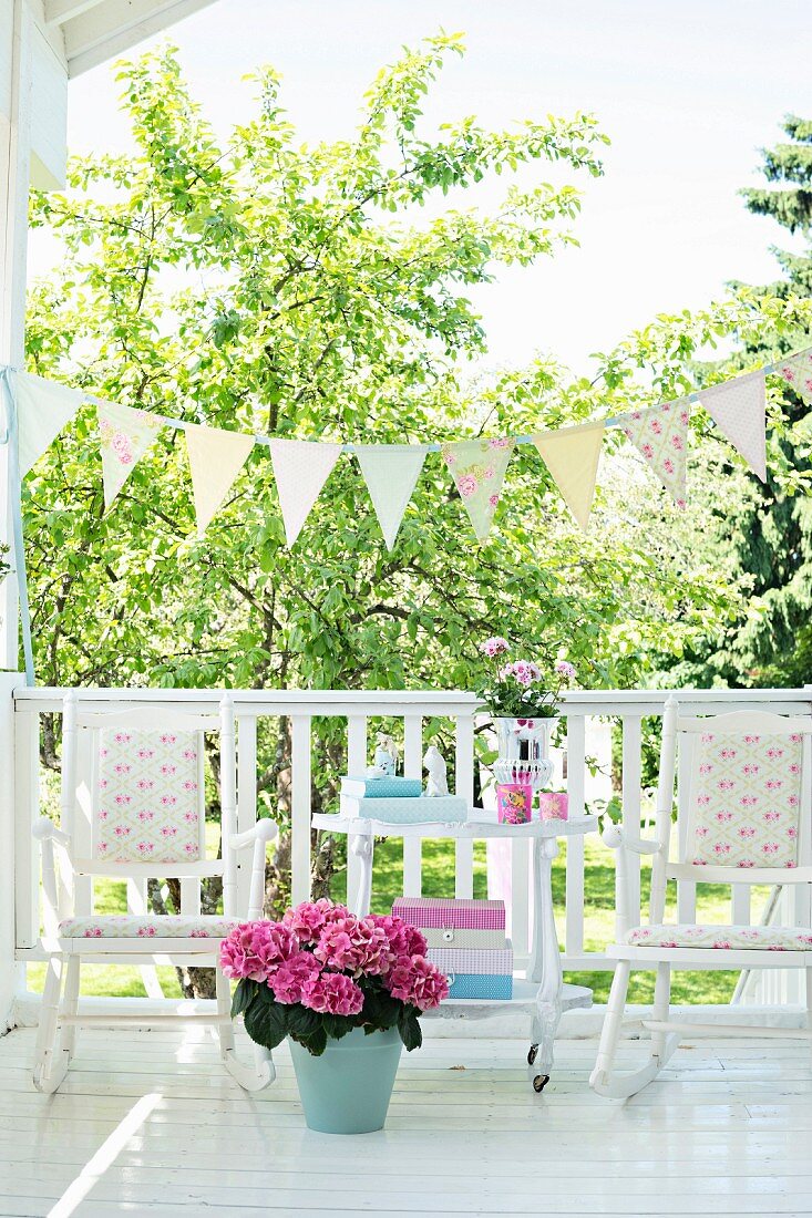 Pinkfarbene Hortensien im Topf vor Schaukelstühle und Tisch, auf Terrasse mit weißem Geländer und Wimpelkette