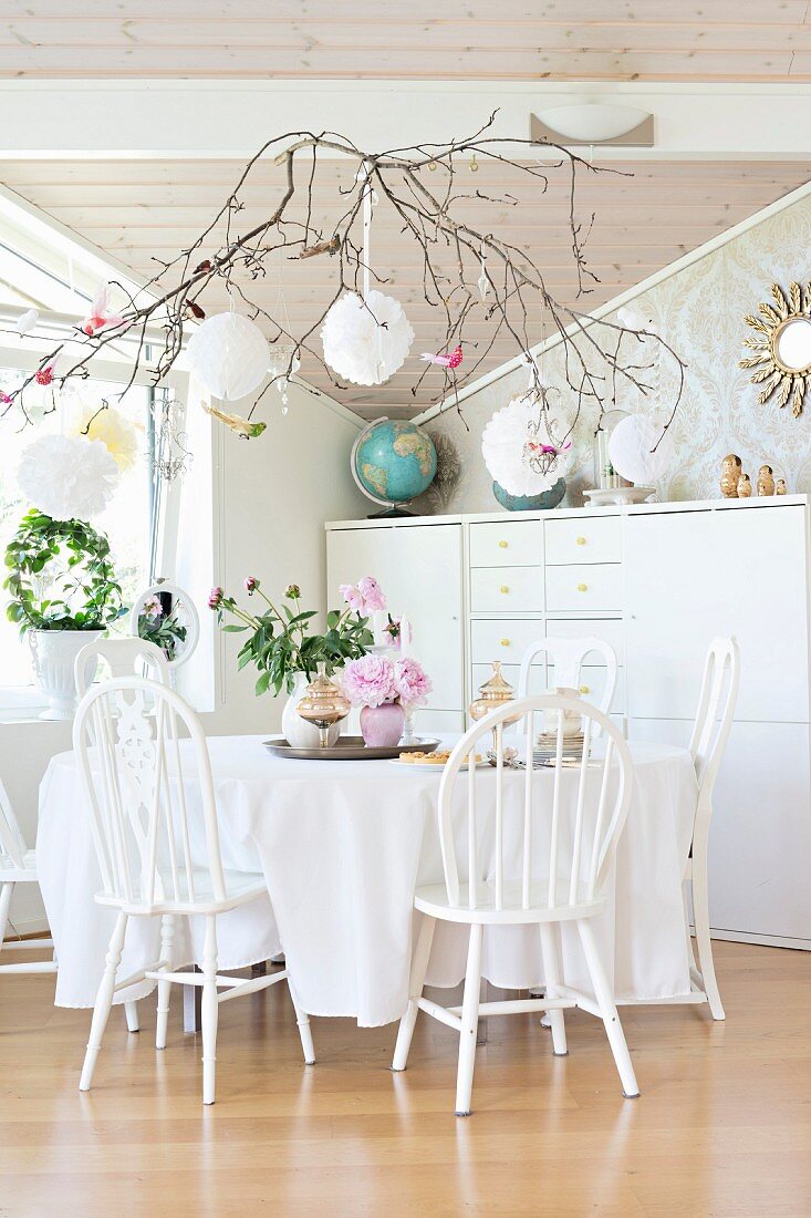 White-painted wooden chairs around table with tablecloth in front of tall sideboard, branches and white ornamental balls suspended from ceiling