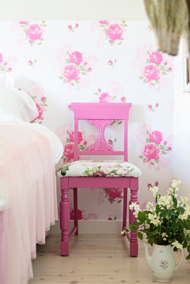 Pink-painted chairs with carved backrest and seat cushions against floral wallpaper next to pot of flowers on floor