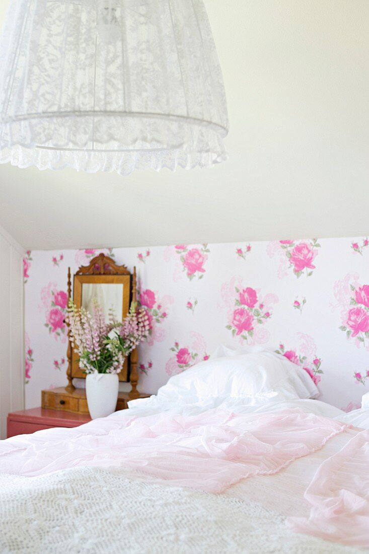 Bedroom with white bedspread on bed, nostalgic floral wallpaper and pendant lamp with lace lampshade