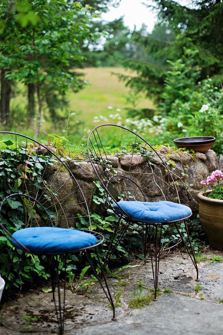 Filigrane Metallstühle mit gebogener Rückenlehne und blaue Sitzpolster, vor niedriger Gartenmauer aus Naturstein