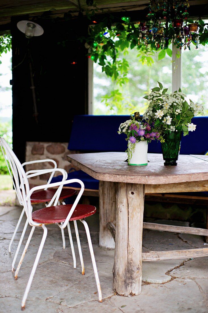 weiße, filigrane Metallstühle vor rustikalem Holztisch, darauf Blumensträusse in Vase, in einer Loggia mit Natursteinboden