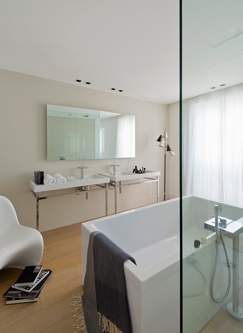 Freistehende weiße Badewanne hinter Glas Trennscheibe, gegenüber zwei Waschtische und Wandspiegel in minimalistischem Designerbad
