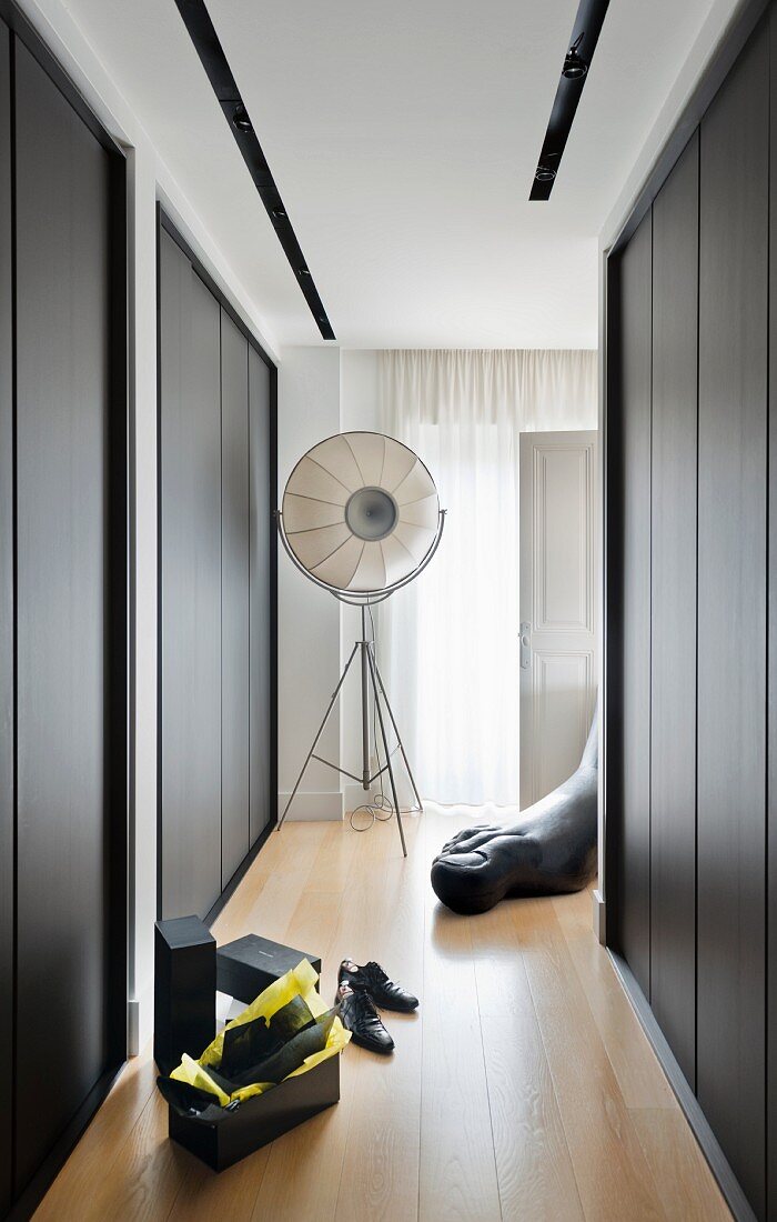 Dressing room with dark fitted wardrobes, shoe boxes and men's shoes on parquet floor; studio lamp and large sculpture of foot in background