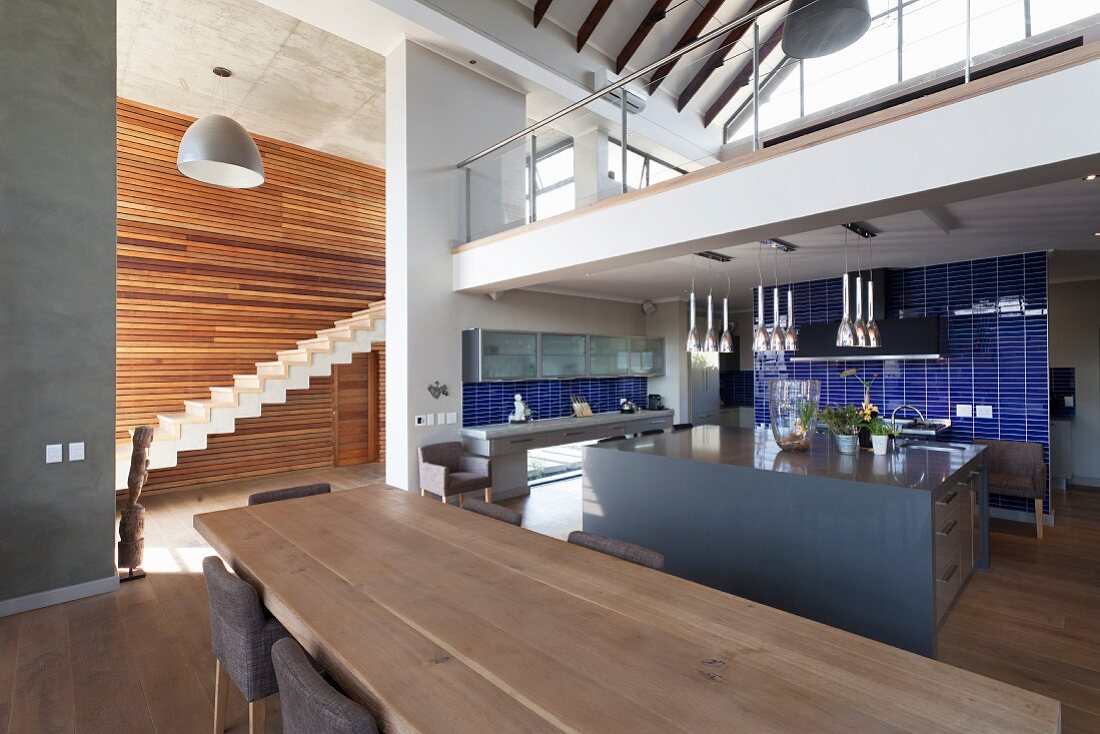 Langer Esstisch aus hellem Holz gegenüber Kücheninsel in offenem Wohnraum, Sichtblicke in Treppenraum mit holzverkleideter Wand und auf Galerie