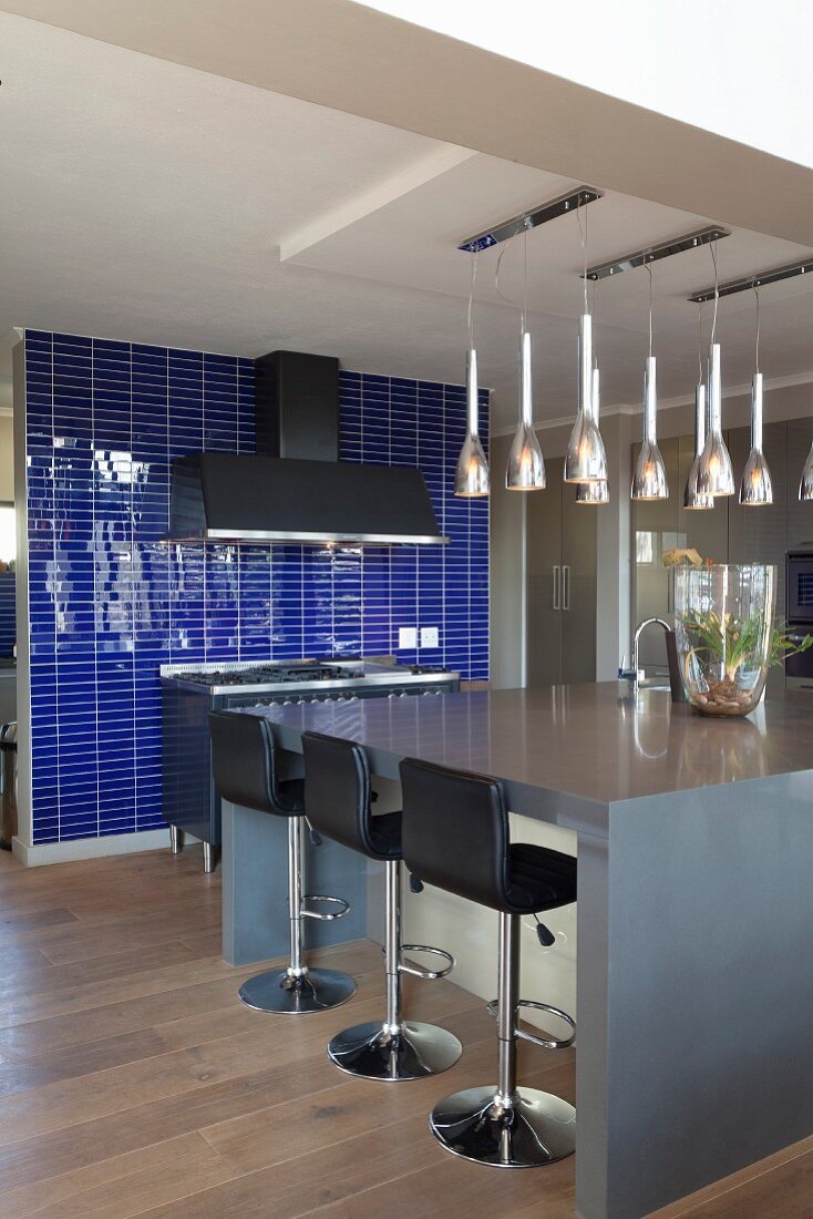 Schwarze lederbezogene Barhocker vor grauer Kücheninsel, darüber mehrere Reihen Hängeleuchten an Decke, im Hintergrund violettblaue Fliesen an Wand
