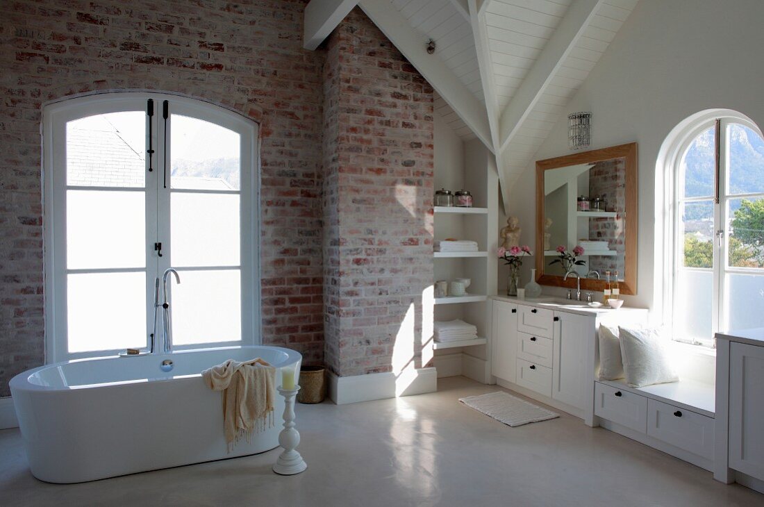 Free-standing bathtub in front of French windows and white fitted cupboards in bathroom with exposed brickwork