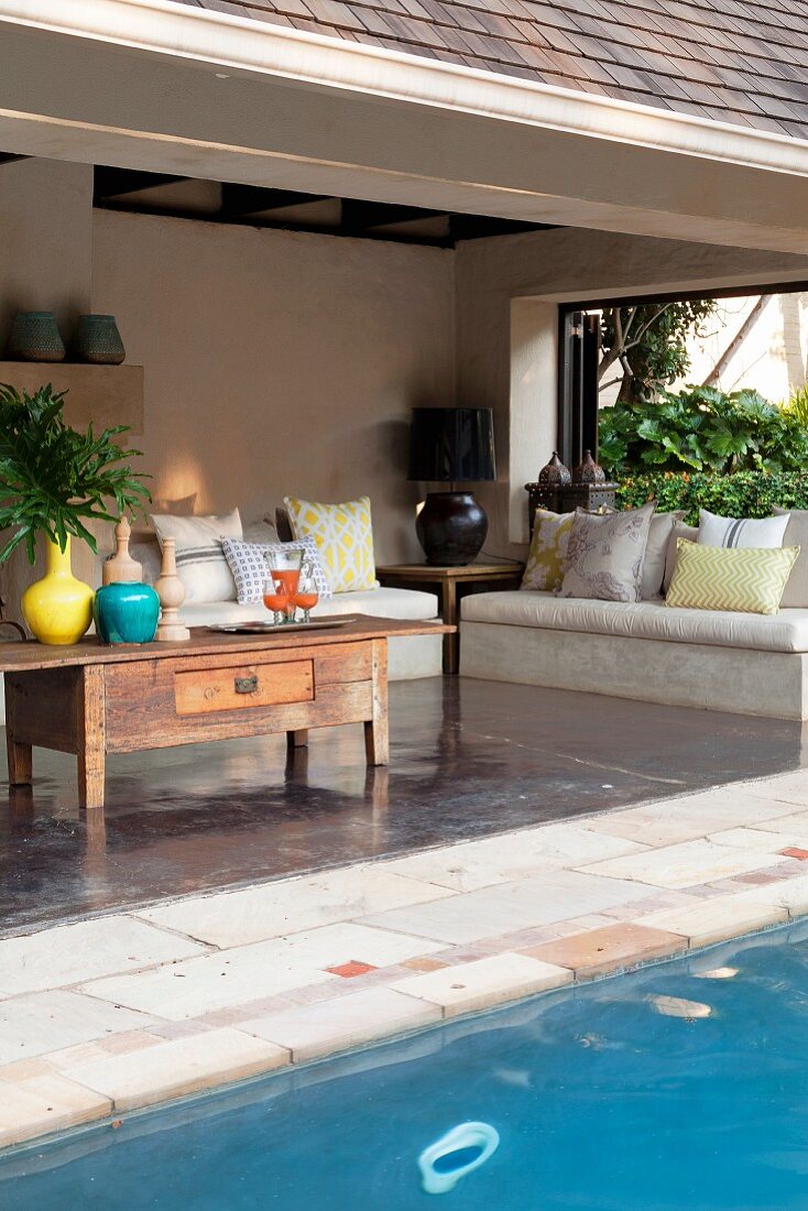 Pool in front of roofed terrace with rustic coffee table; concrete bench with cushions in background