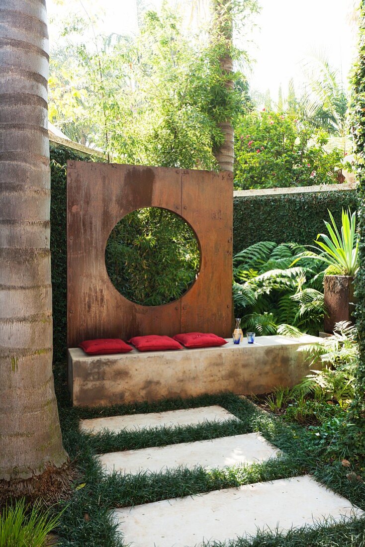 Steinplatten auf Wiese vor Sitzbank mit roten Kissen und aufgestellte Wand aus rostigem Metall mit kreisförmigem Ausschnitt in tropischem Garten