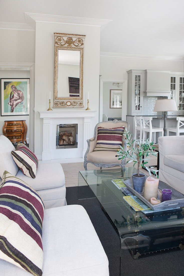 Pale sofa with striped scatter cushions, curved glass coffee table and Rococo armchair in front of fireplace in open-plan interior