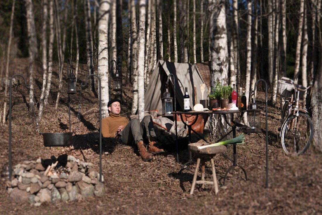 Campfire, picnic, table, tent and bicycle in autumn woods; man relaxing with back against tree trunk