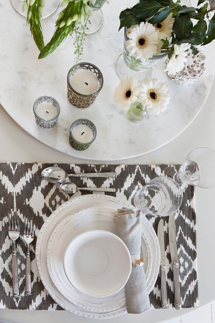 Place setting with white crockery and patterned table mat next to tealight holders and vases of flowers on round dish