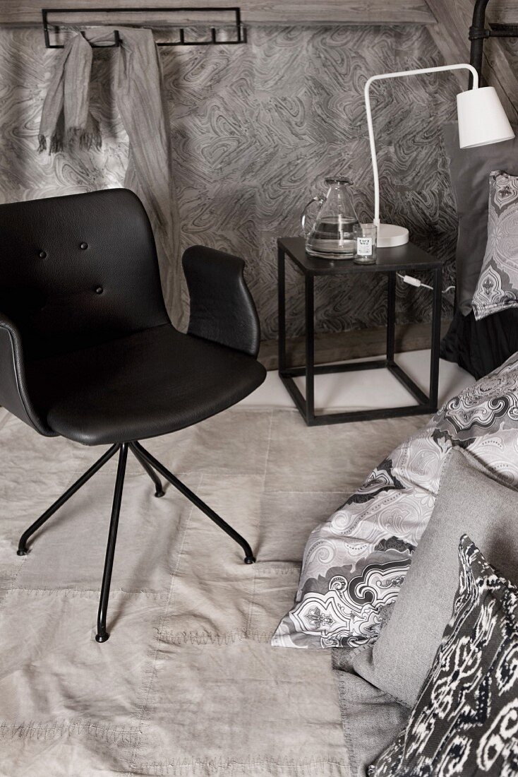 Designerstuhl neben Kissen am Boden, kubischer Beistelltisch mit weisser Nachttischleuchte und grau gemusterter Tapete