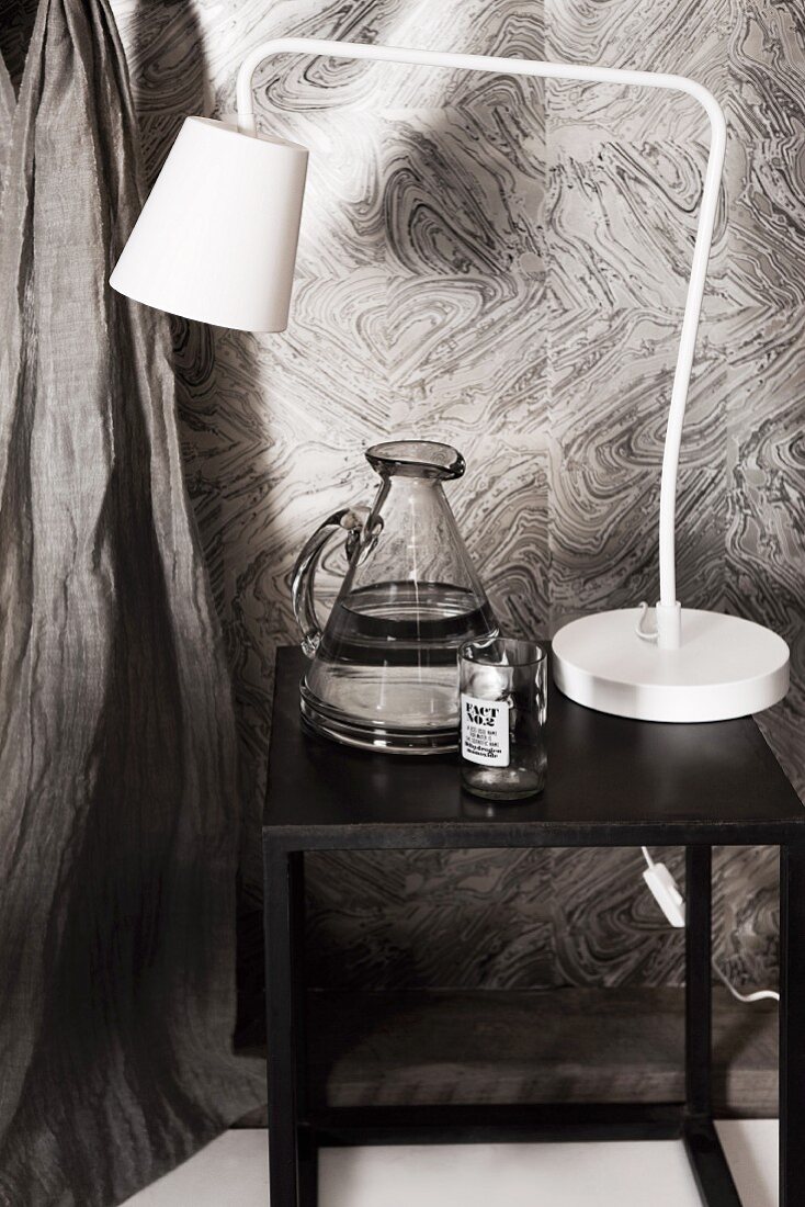 White, retro table lamp and jug of water on black side table against grey-patterned wallpaper