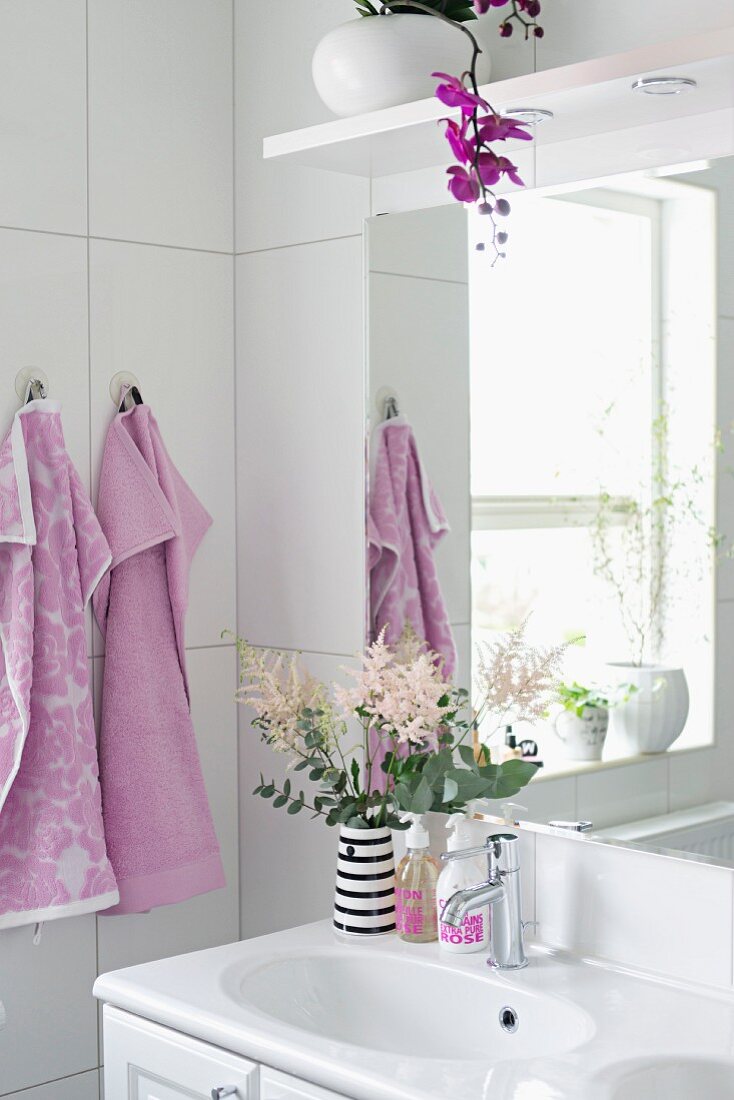 Waschbecken mit Blumendeko, Orchideenblüte über Spiege, seitlich fliederfarbene Handtücher in weißem Badezimmer