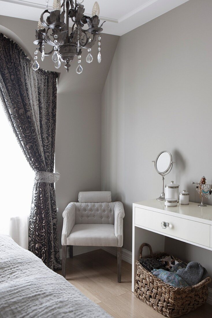 Upholstered chair next to gathered curtain in corner of bedroom painted pale grey; modern console table with cosmetic mirror to one side