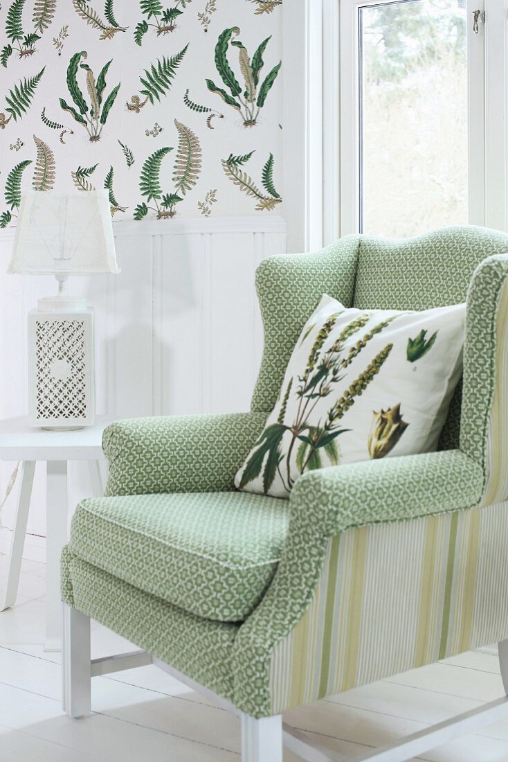 Sessel mit hellgrün gemustertem Bezug und Kissen neben weißem Beistelltisch mit Tischleuchte, an Wand Tapete mit Farnmotiven