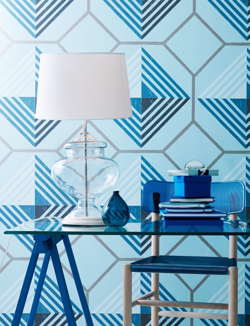 Tischlampe mit weißem Schirm auf Glastisch, vor Tapete mit geometrischem Muster in verschiedenen Blautönen