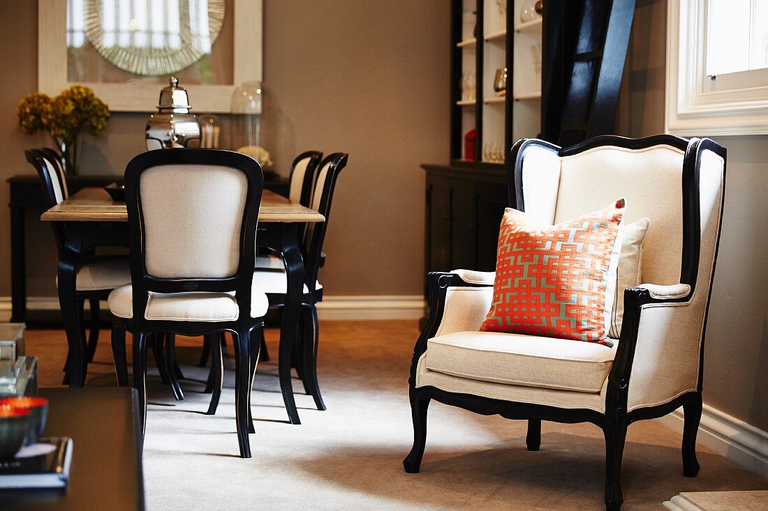 White covered armchair with black wooden frame and scatter cushions next to dining set with chairs upholstered in similar style in traditional interior with walls painted pale brown