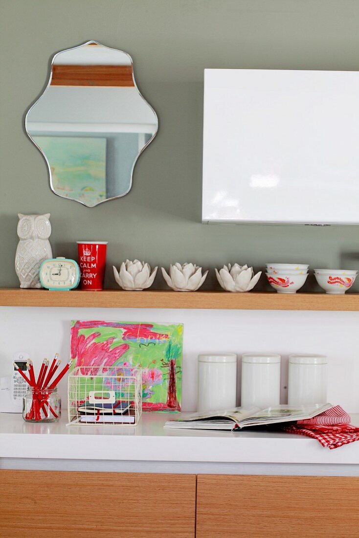 Kitchen counter with white worksurface, storage jars next to child's drawing and various ceramic bowls on white wooden shelf on grey-painted wall