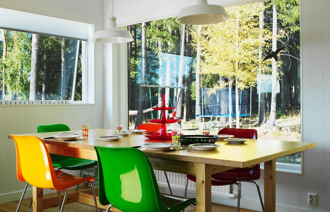 Bunte Schalenstühle um einen Holztisch in Raumecke; Blick in den Wald durch Panoramafenster