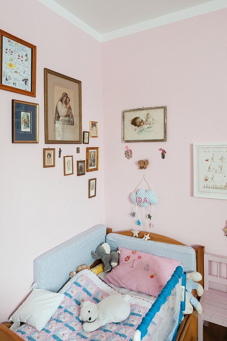 Kinderbett mit Stofftieren in Zimmerecke, an rosa getönten Wänden gerahmte Bilder