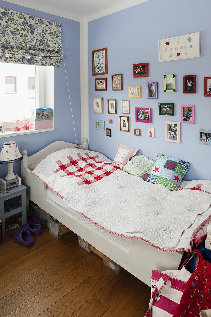Single bed with white-painted wooden frame below framed pictures on lilac wall in corner of child's bedroom