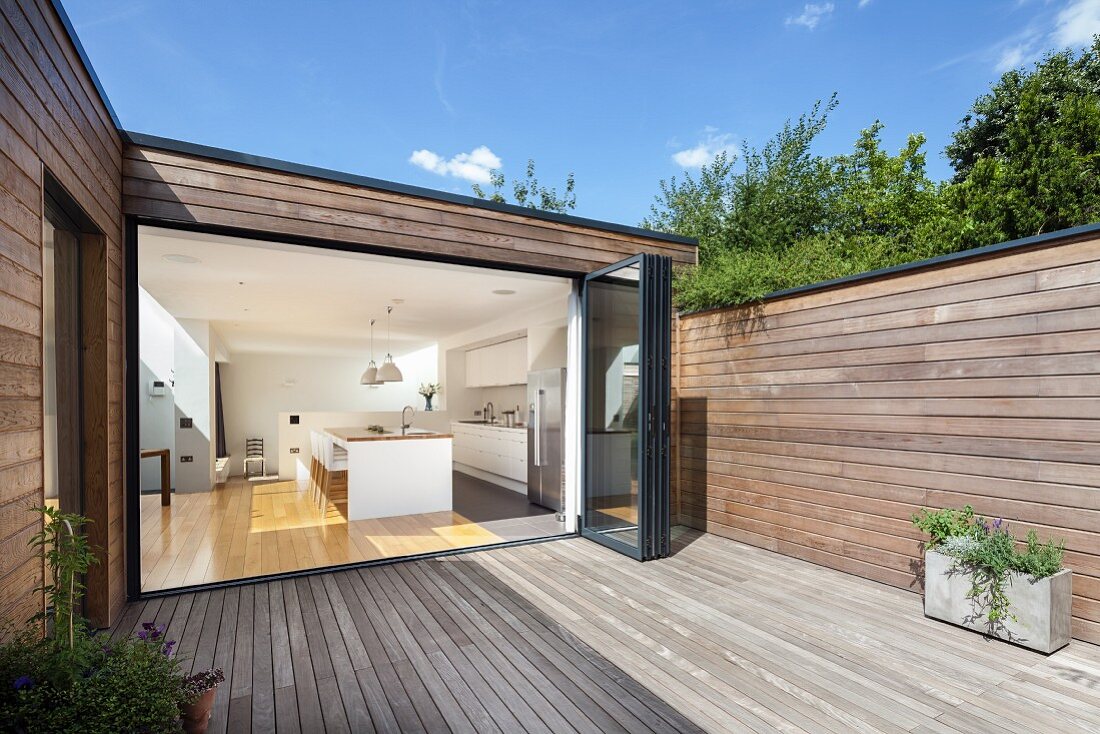 Wooden deck of contemporary house with wood-clad facade; view into kitchen through open folding door