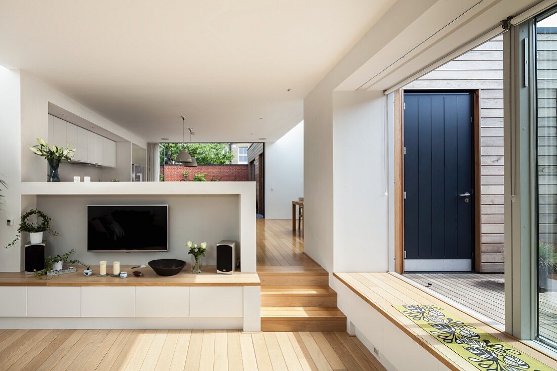 Open-plan interior with TV on half-height wall, wooden steps leading to dining area and open sliding doors leading to terrace