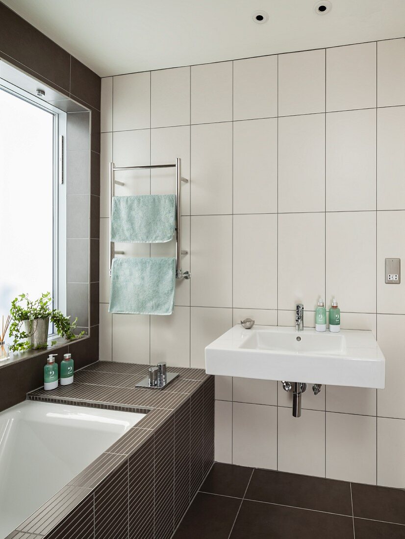 Waschbecken an Wand mit weissen, hochformatigen Fliesen, seitlich eingebaute Badewanne am Fenster im Designerbad
