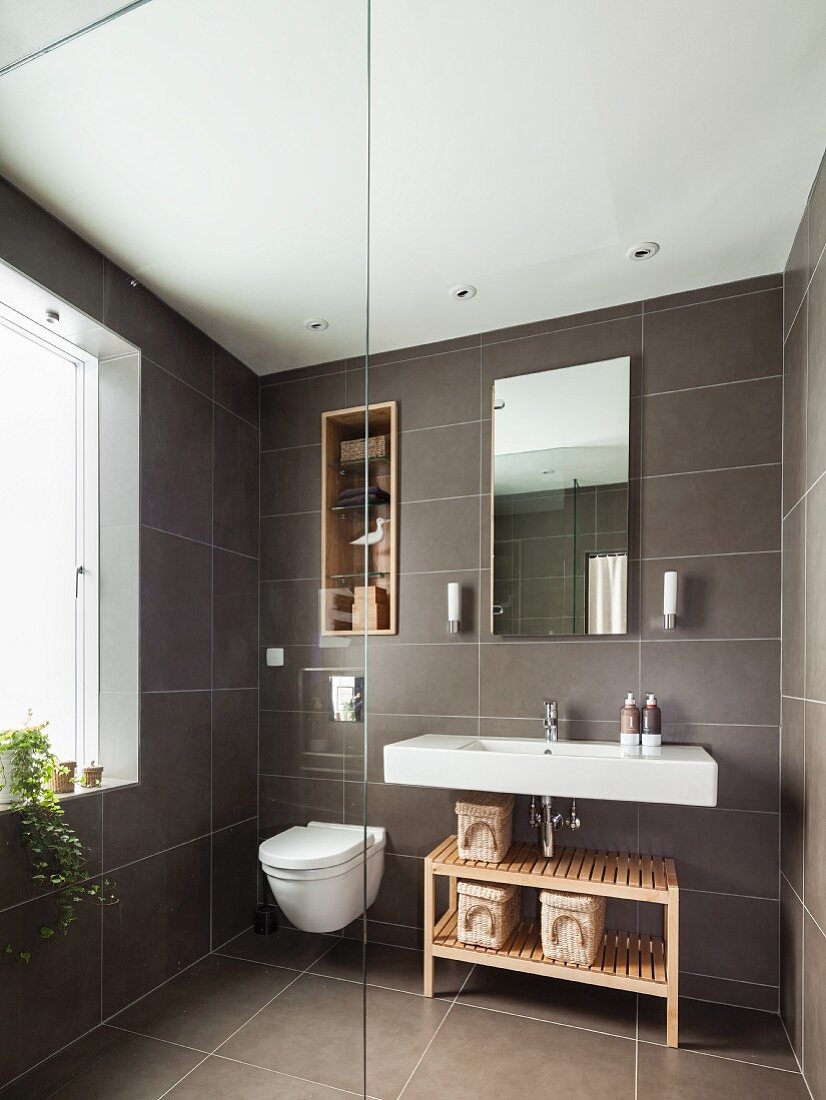 Grey-tiled bathroom with shelves in niche above toilet next to sink and wooden shelves; glass partition in foreground