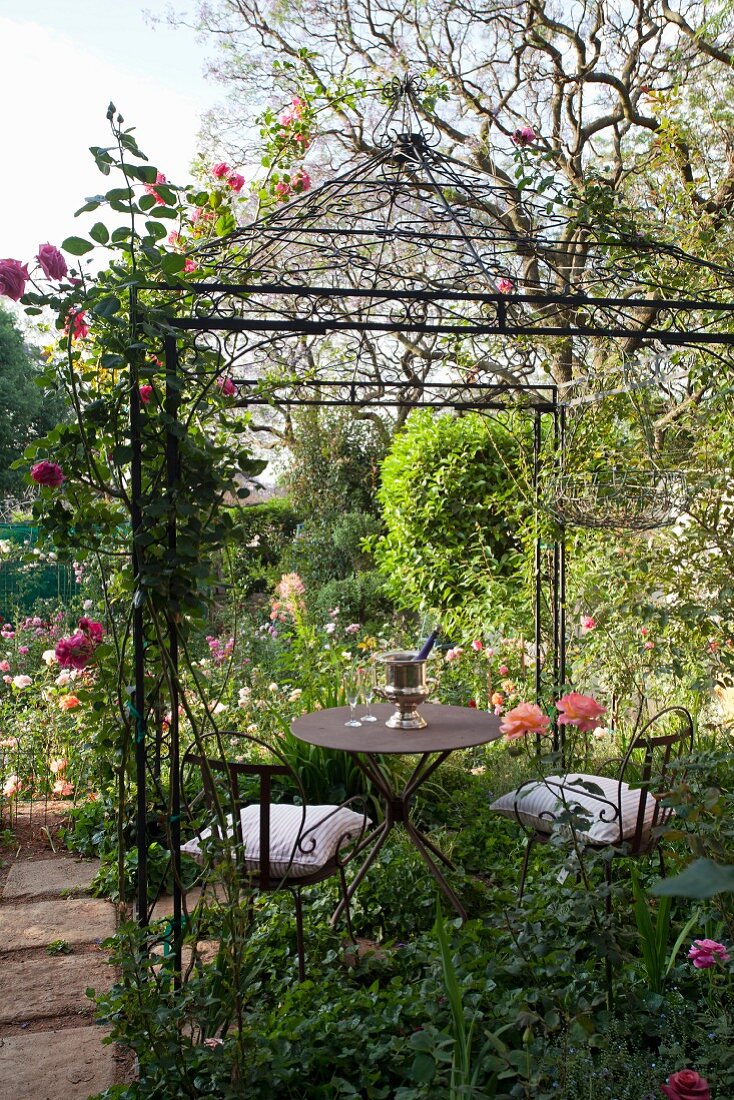 Sitzplatz mit Metallmöbeln in rosenberankter idyllischer Gartenlaube