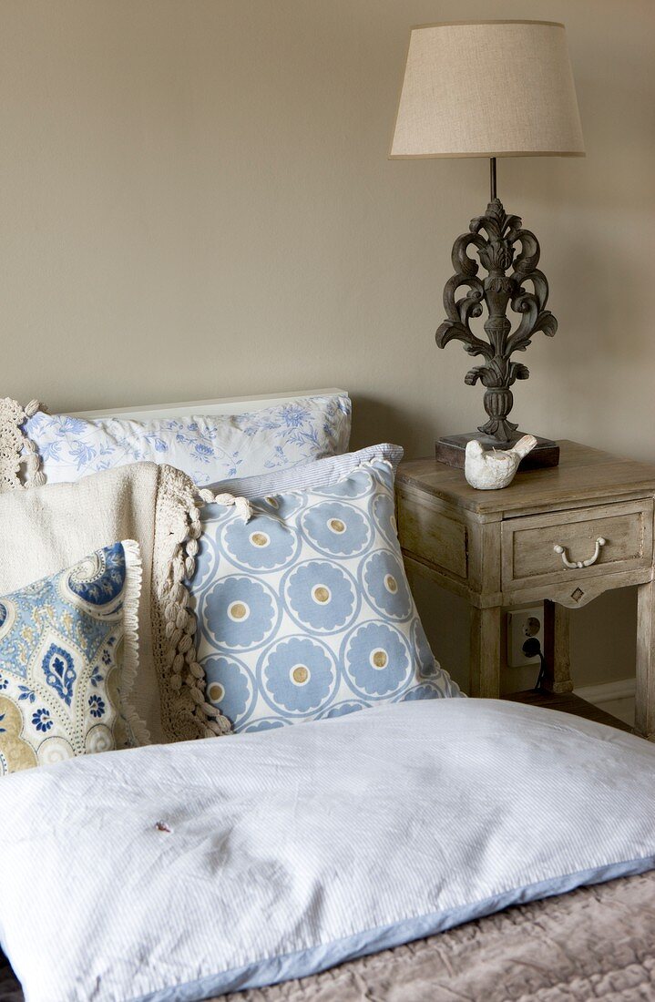 Tischleuchte mit klassischem Lampenfuss auf Nachtkästchen im Antikstil und zartblau gemusterte Kissenparade auf dem Bett