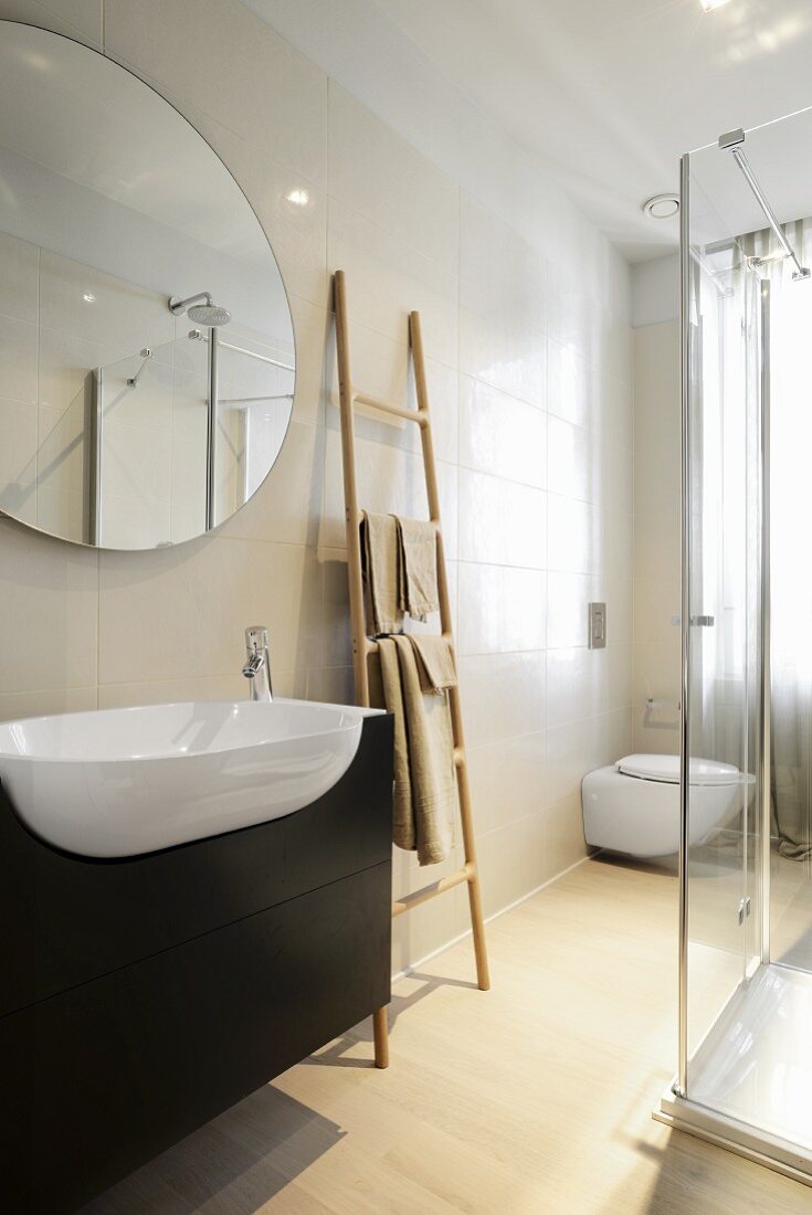 Trogartiges Waschbecken mit Unterbau aus dunklem Holz, ovaler Spiegel an gefliester Wand, daneben leiterartiger Handtuchhalter, gegnüber teilweise sichtbare Glas Duschkabine