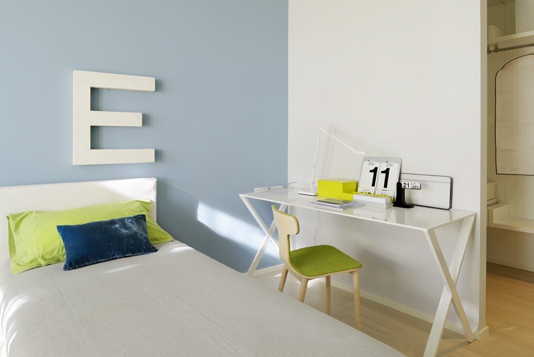 Einzelbett mit Kopfteil und grossformatiger Buchstabe an pastell lila Wand, neben weißem Metalltisch mit Stuhl vor Raumteiler