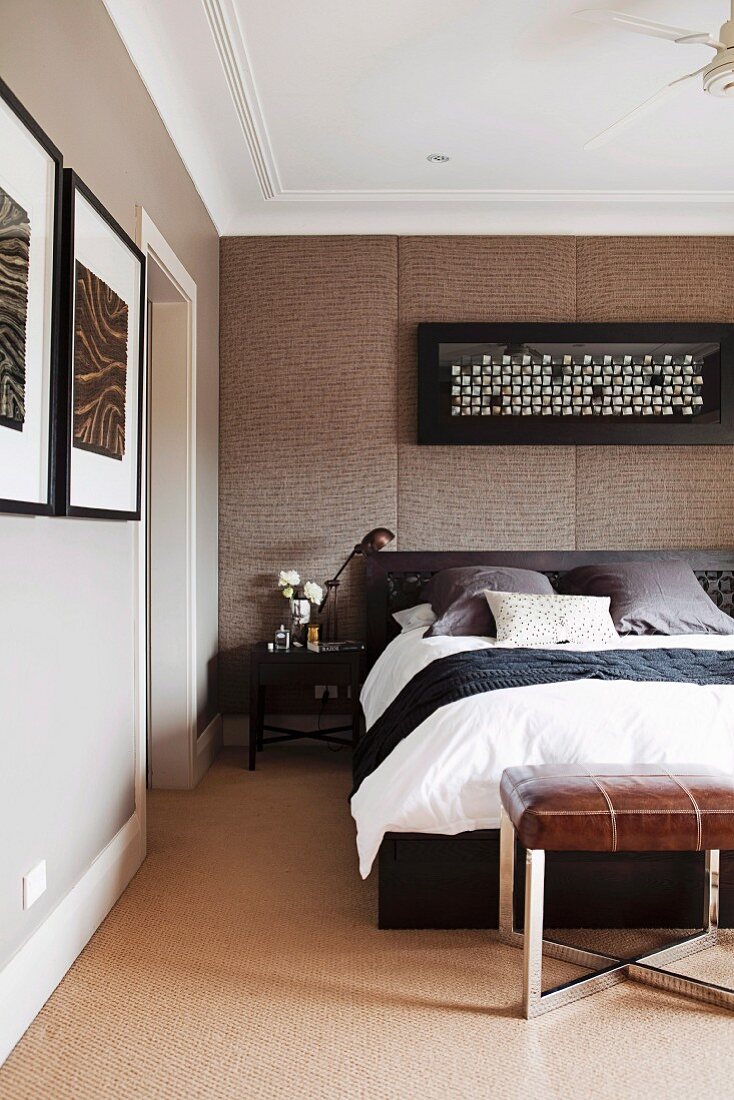 Doppelbett mit Kopfteil vor gepolsterter Wand, unter gerahmtem Bild, am Bettende gepolsterter Hocker mit Lederbezug im Schlafzimmer